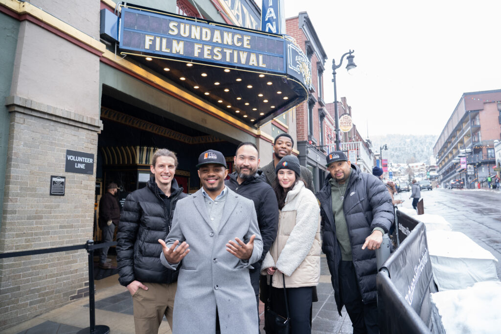 Six people wearing winter wear standing under a theater marquee that reads 'Sundance Film Festival'.
