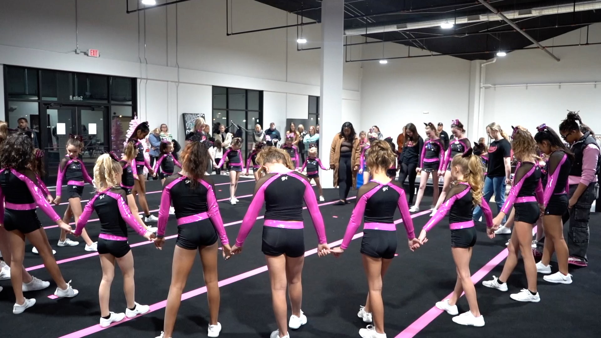 About two dozen young people hold hands with their heads bowed in a circle. They wear pink and black dance wear and white sneakers.