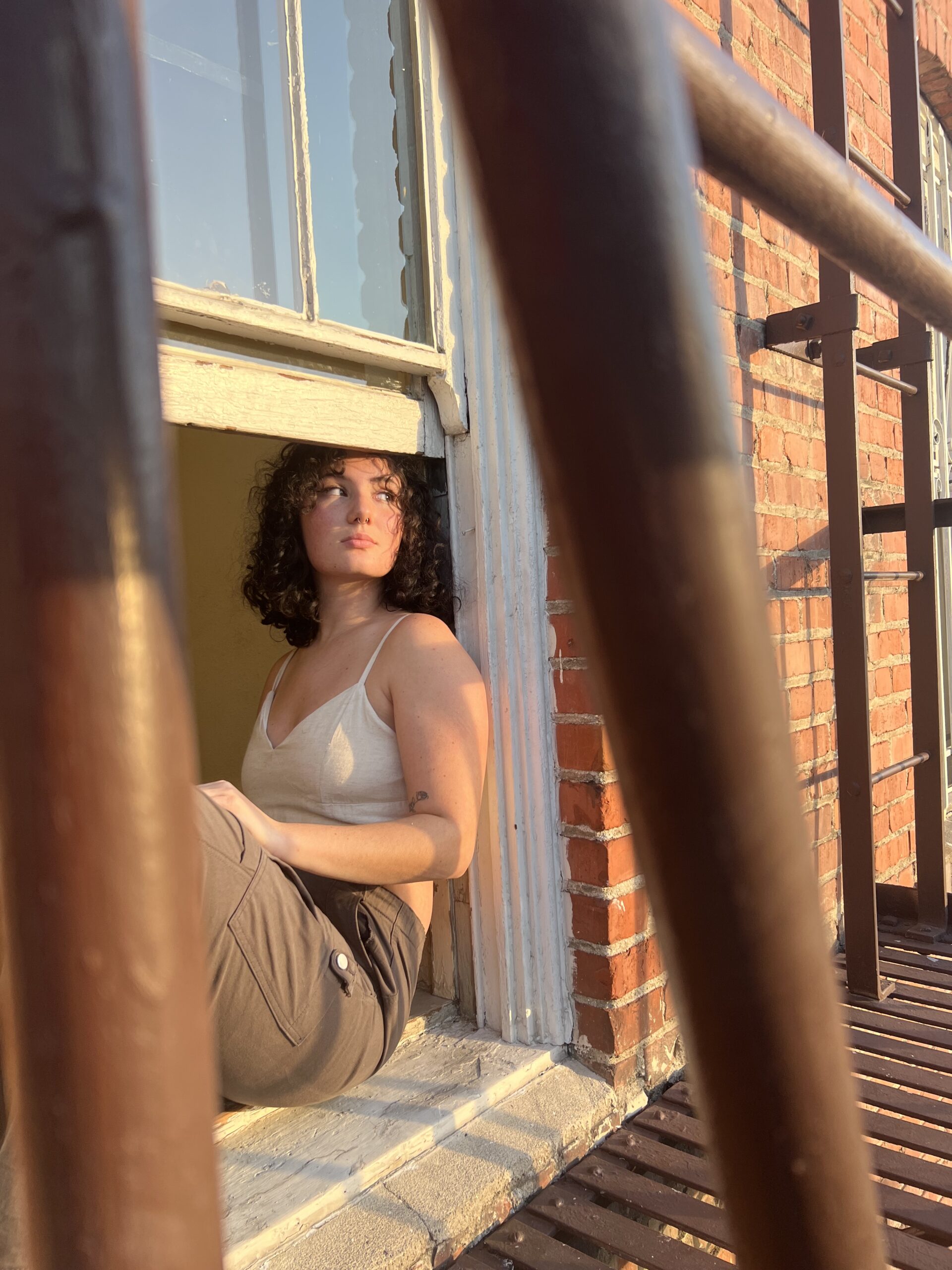 A person of light skin tone and curly dark hair sitting on a window sill by a fire escape and looking into the distance.