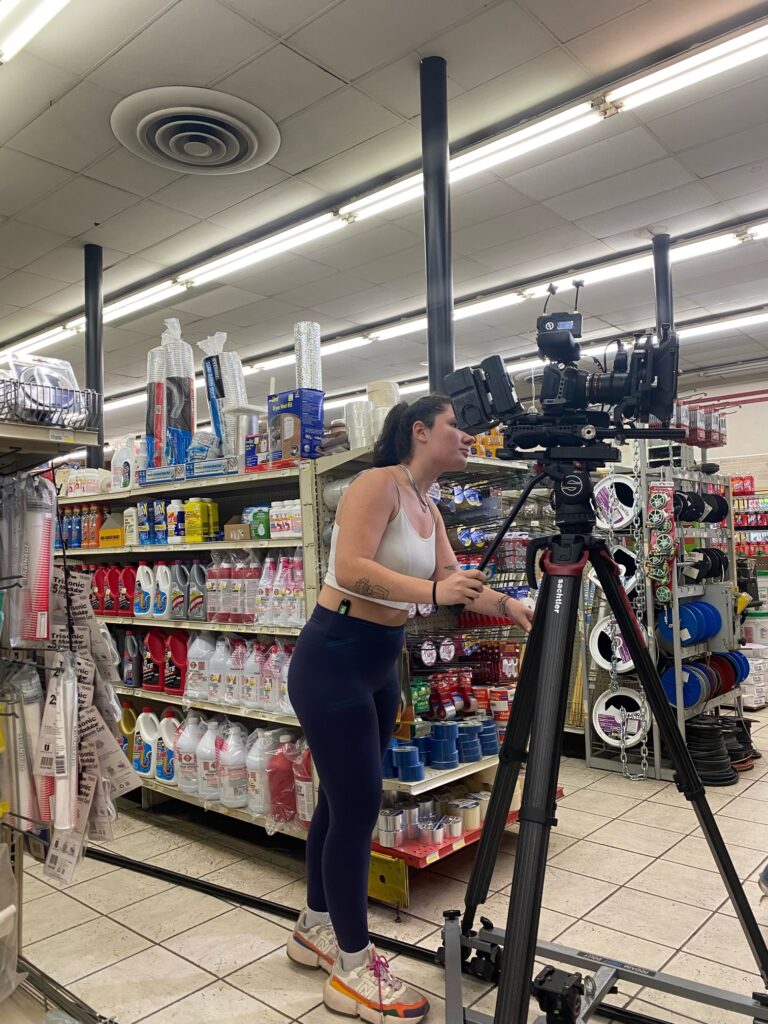 A person of light skin tone standing by a video camera on a tall tripod at a hardware store. They are looking through the camera's monitor and resting their hands on the tripod.