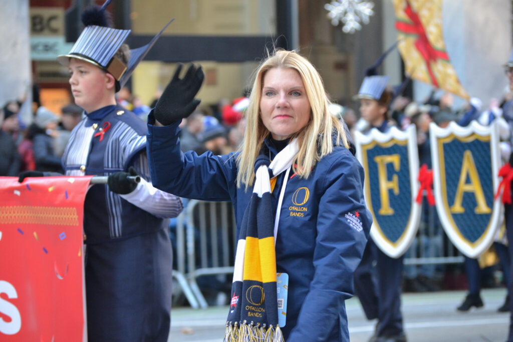 A person of light skin tone and blonde hair waves as part of a street parade. There is a young adult beside them wearing a navy blue marching band uniform.
