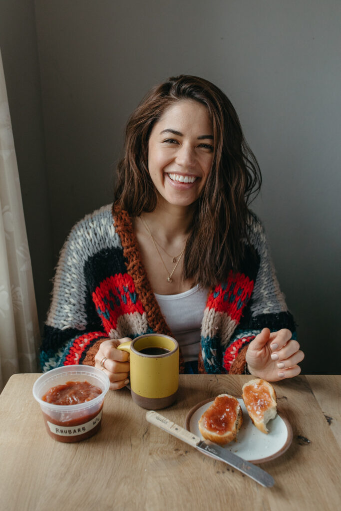 A smiling person with long dark hair sits at a table holding a yellow mug of coffee. There is a plate with two pieces of toast with jam and a container of jam labeled "rhubarb."