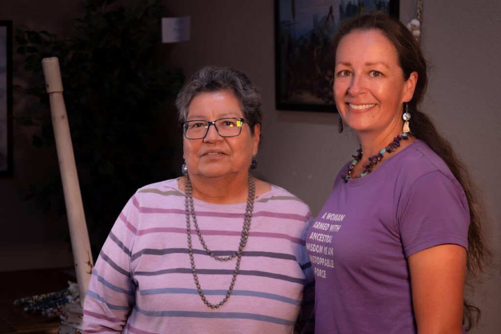 Two people stand close together smiling at the camera. One wears two long beaded necklaces, glasses, and has cropped gray hair. The other is wearing a beaded necklace, beaded earrings, and has a long dark braid down their back.