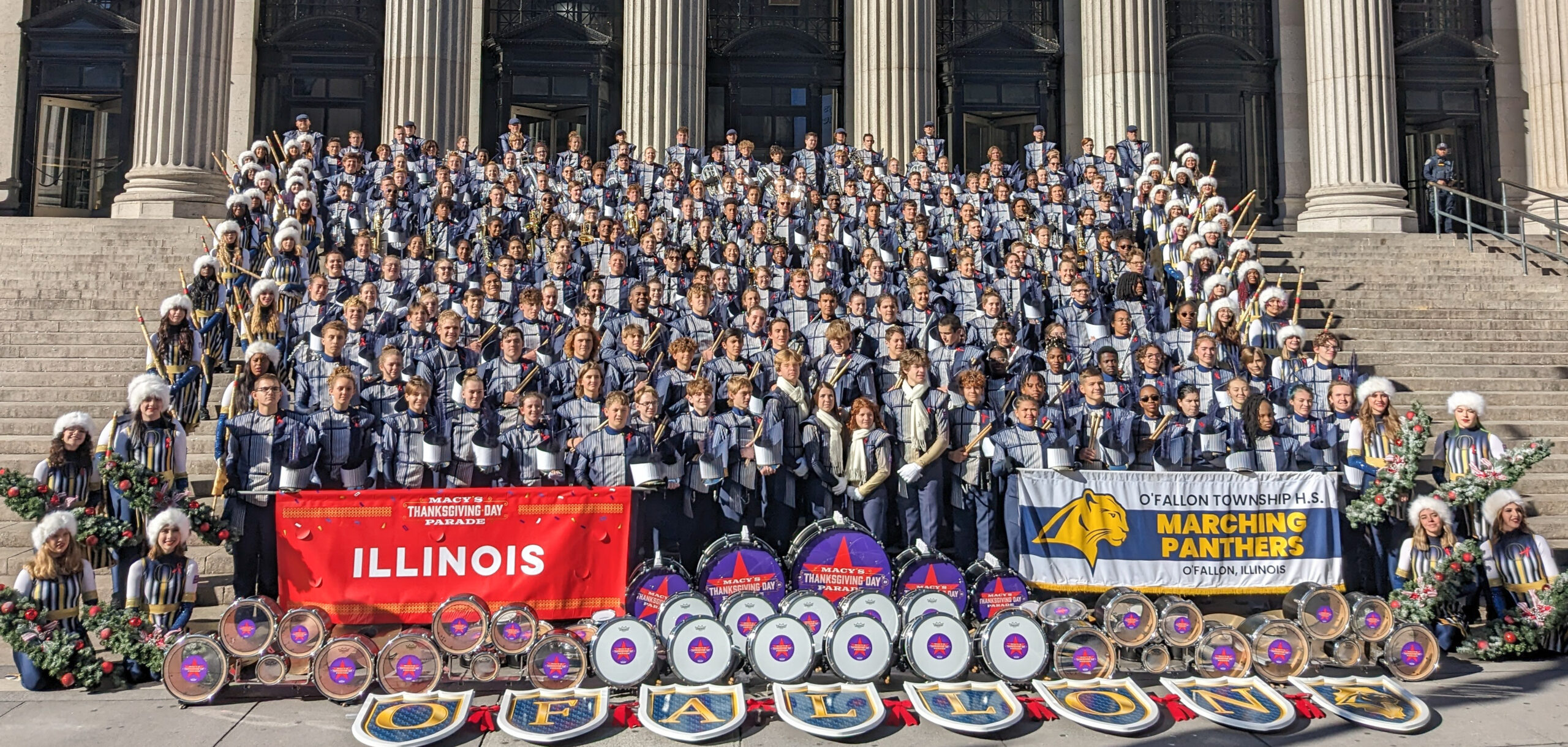 A large group of young adults standing on a tall set of stairs. They are all wearing navy blue and white marching band uniforms. There is signage in front of them that read 'O'Fallon'