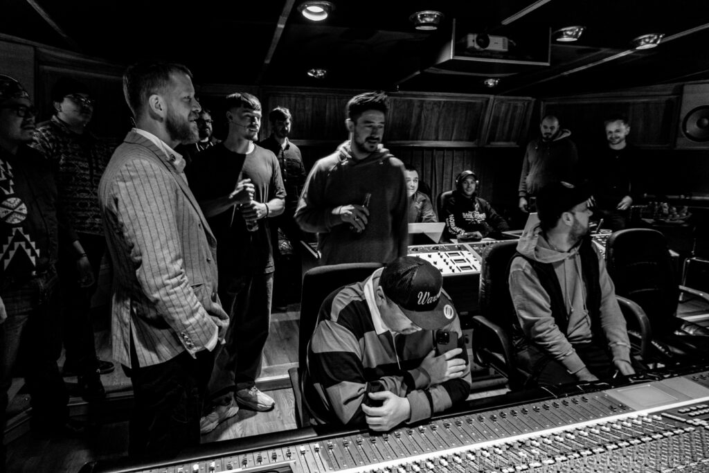 A group of people in a recording studio. Some are standing while others are seated by recording and mixing panels.