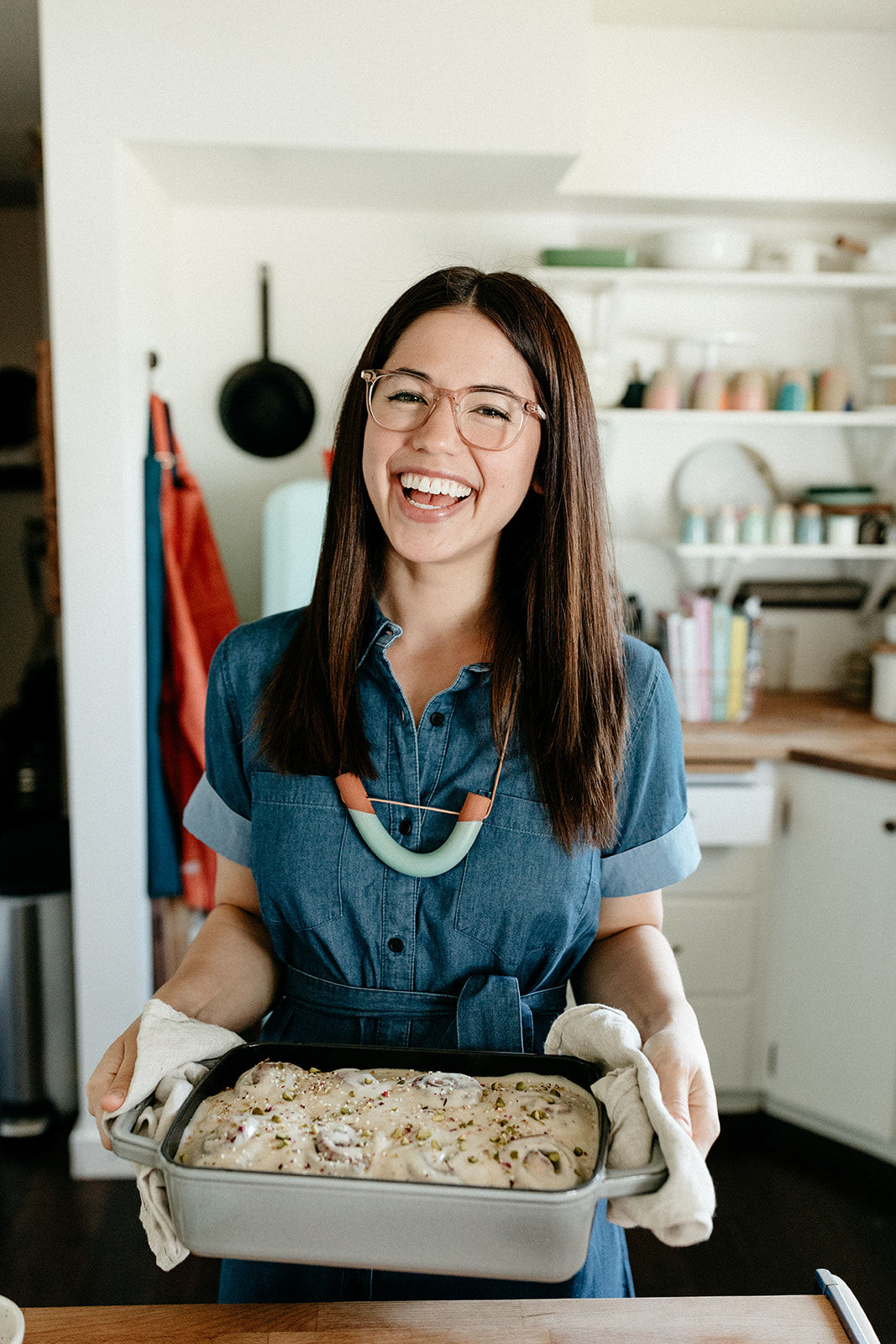 A smiling person wearing a blue dress, brightly colored necklace, glasses, and oven mits, holds a baking tin filled with a white subtance.