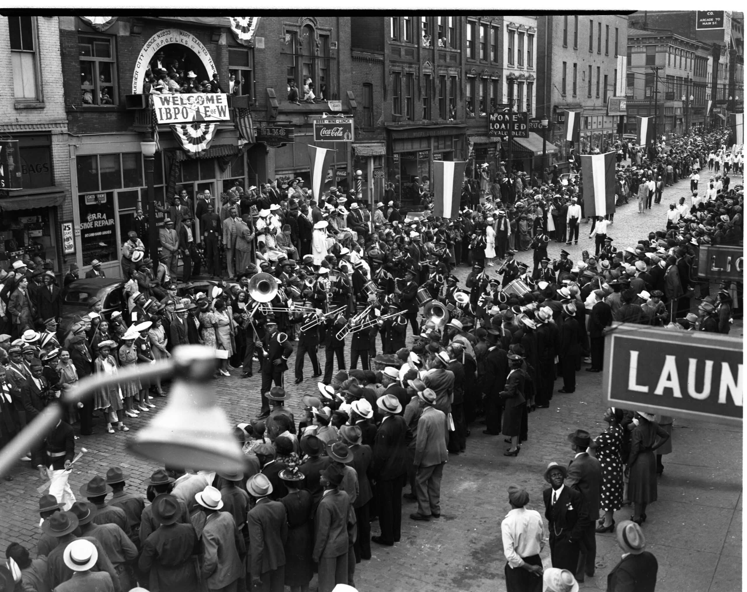 An archival black and white photo of a strett parade with a brass band and crowds all over the street.
