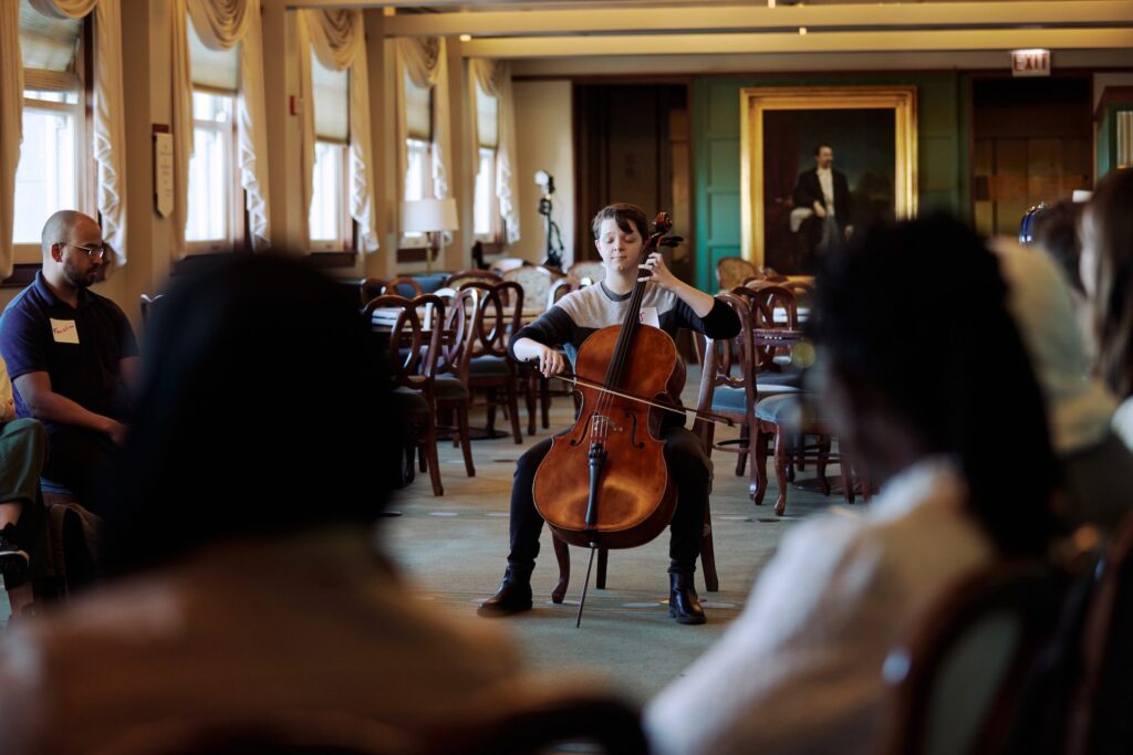 A person plays the cello with their eyes closed in the middle of a room scattered with tables and chairs. In front of them. is an audience with their backs to the cameras.