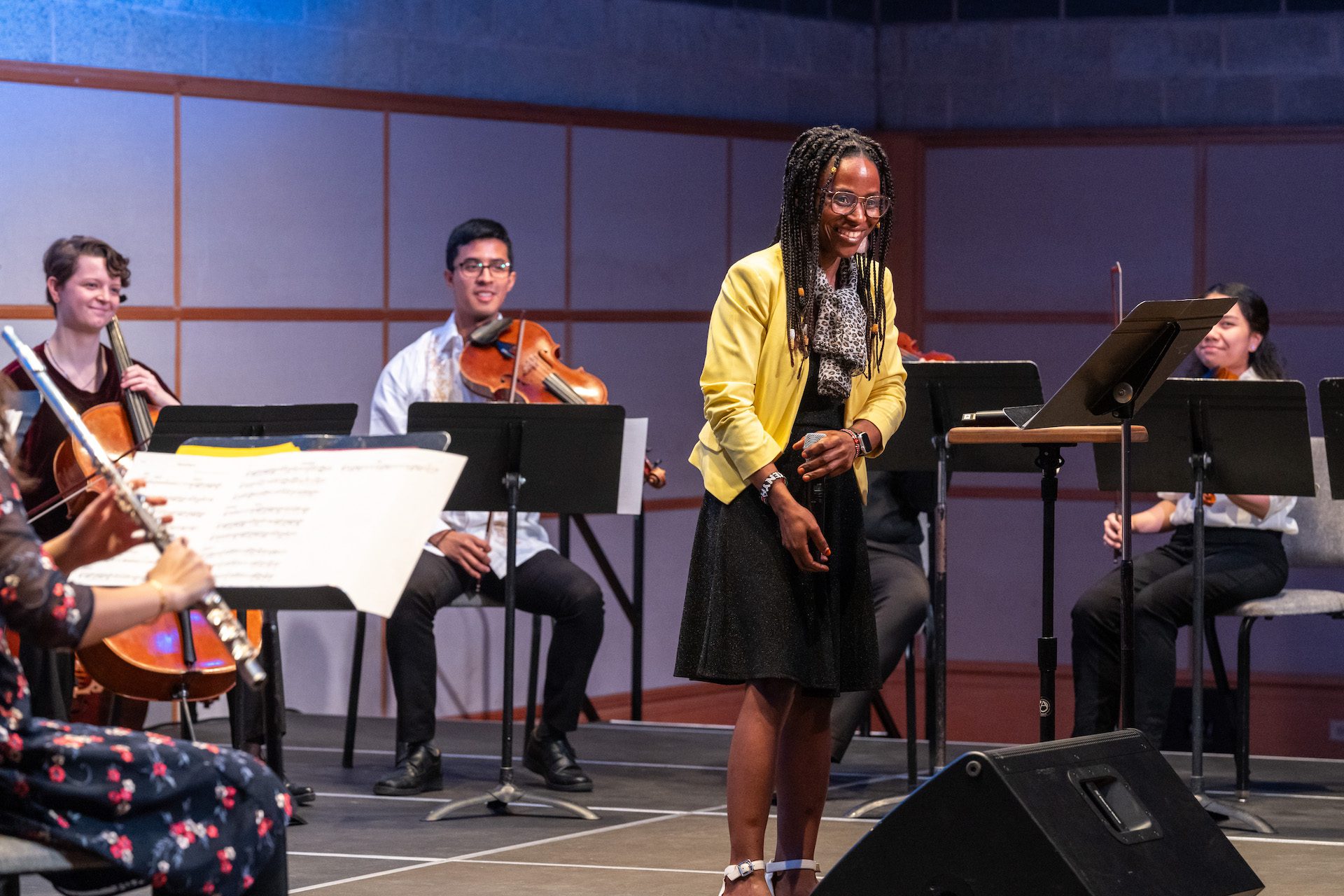 A person in a bright yellow jacket stands smiling in the center of a half-circle of seated musicians. The musicians hold violins, a cello, and a flute.