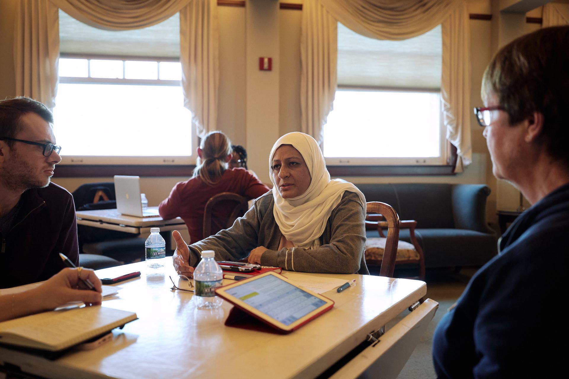 Three people seated at a table -- one of them, wearing a white headscarf, is talking as the two others look on. In front of them are open notebooks and an iPad.