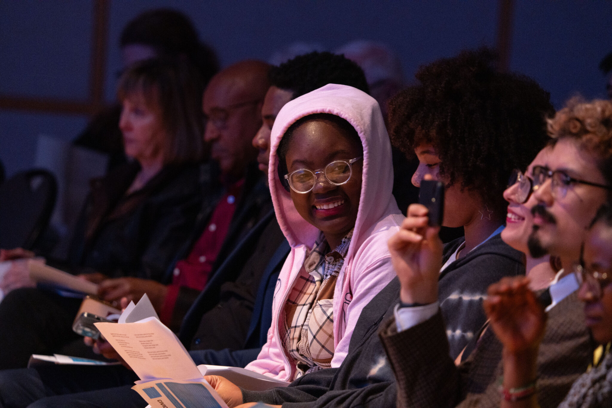 A row of people are seated close together. Most of them look ahead smiling. One person wearing a bright pink hoodie over their head and glasses is turning to smile brightly at someone sitting next to them.