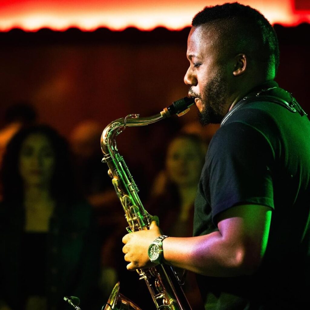 A person of dark skin tone wearing a dark tshirt and playing the saxophone.