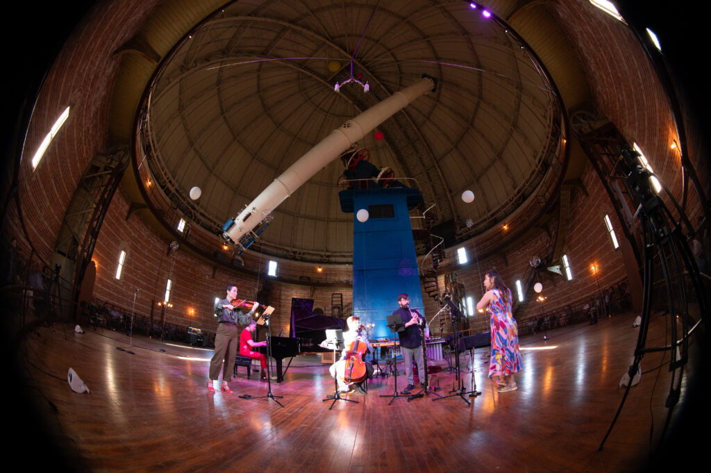 Five musicians perform in a domed room under a large telescope. They are playing various instruments.