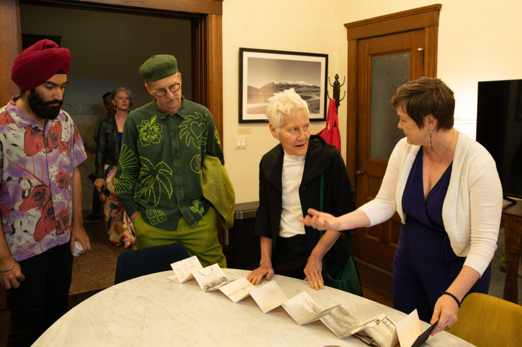 Four people stand beside a marble table as they gesture and talk to each other. There is an accordion-folded piece of paper on the table.