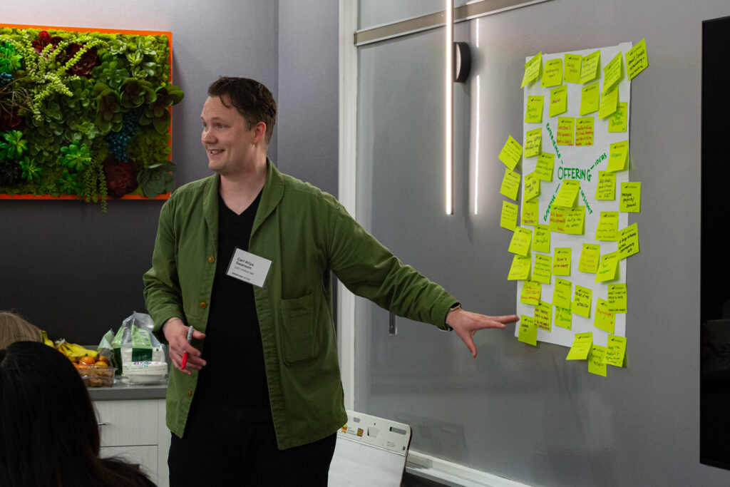 A person of light skin tone and dark short hair wearing a green jacket stands by a wall with bright post-in notes and gestures.