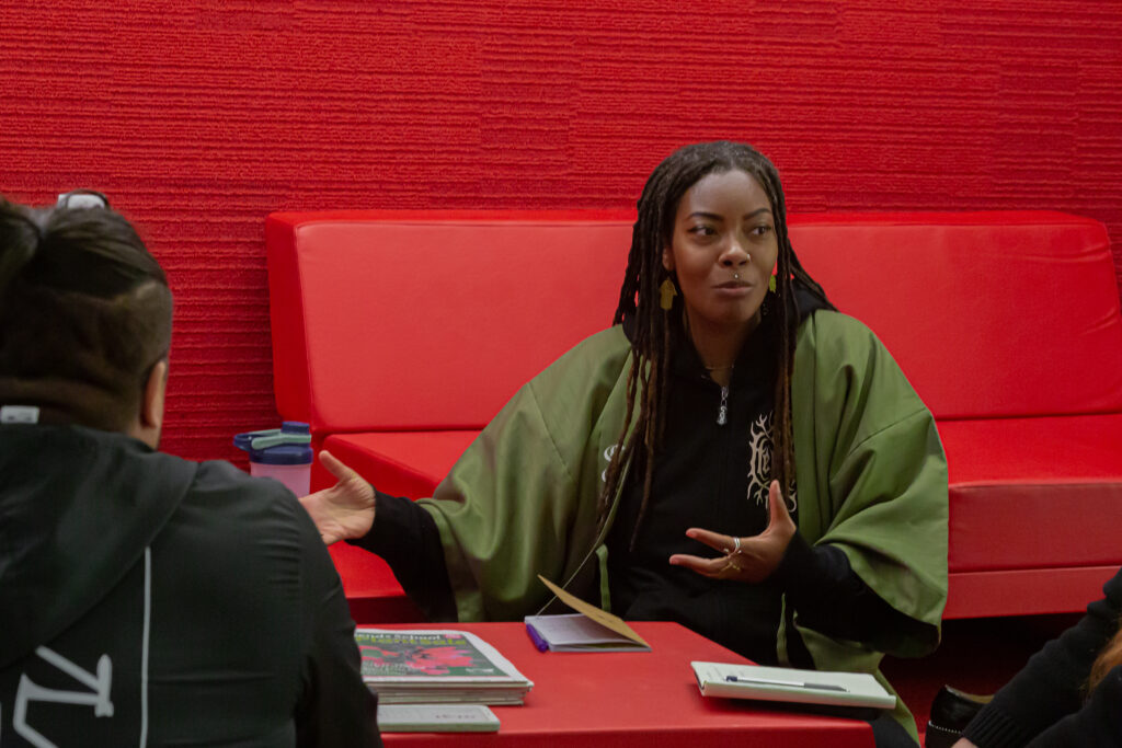 A person of dark skin tone and dark, long dreadlocks wearing a green jacket gestures and talks to people. They are in an area with red walls, a red couch, and a red table.