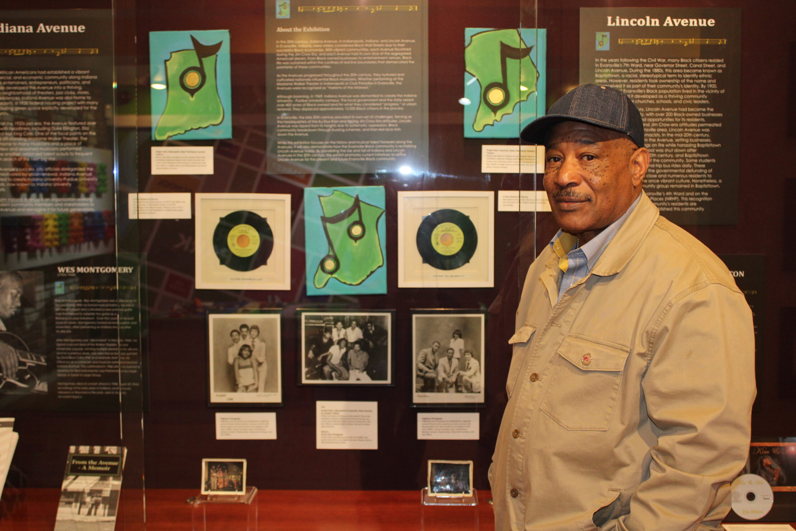 A person of medium dark skin tone wearing a khaki colored jacket and a denim baseball hat stands in from of a museum display case.