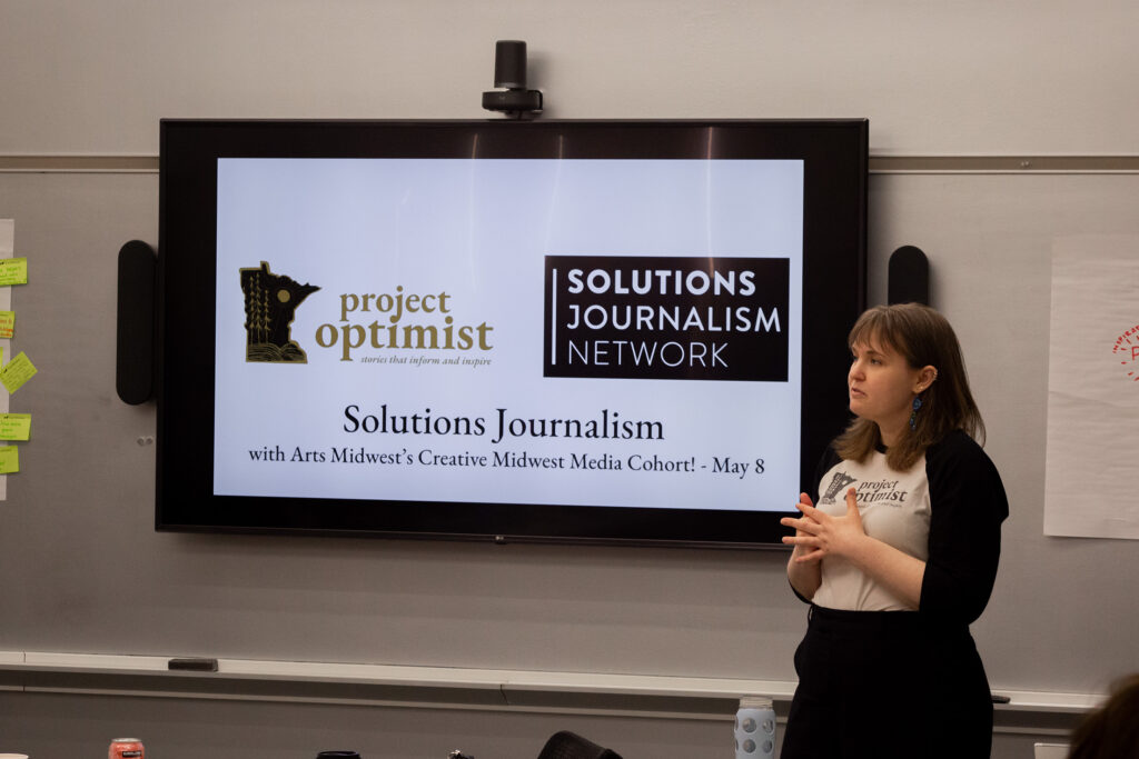 A person of light skin tone and dark medium length hair stands by a television screen with a slide that has logos of Project Optimist and Solutions Journalism Network.