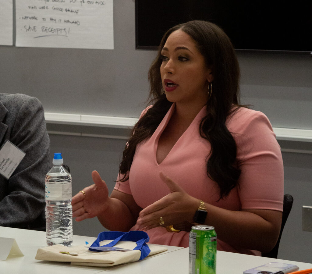 A person of medium dark skin tone and long dark hair wearing a peach dress sits at a table. They are gesturing with their hands as they speak.