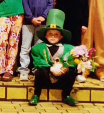 A small child in a green leprechaun suit and hat sits on a step painted to look like the Yellow Brick Road.