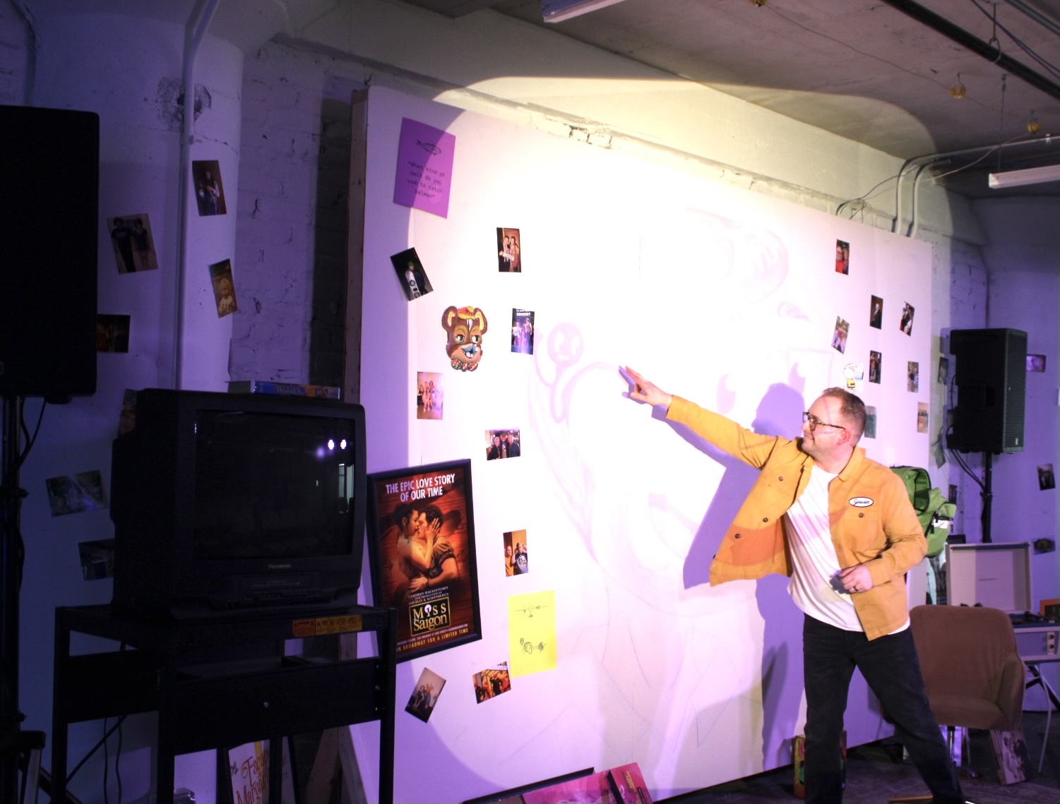 A person in a bright yellow jacket smiles and points to a spot on a large white wall lined with photos and a promotional poster for Miss Saigon. There is what appears to be a cartoon face of a large duck projected on the wall with a light projector.