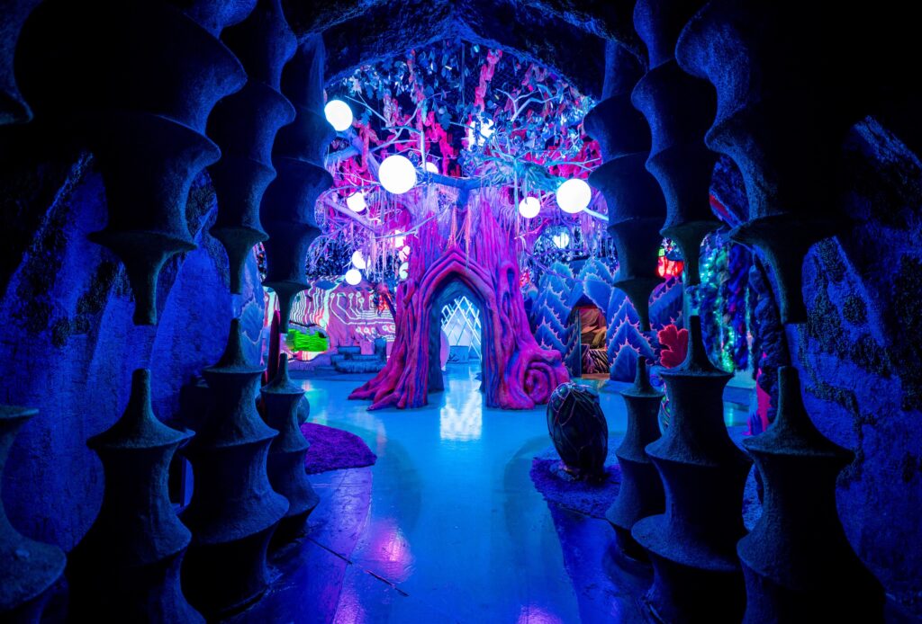 An art installation depicting a room with glow-in-dark decorative accents and light fixtures. There is a walkway through a tree stump-like work.