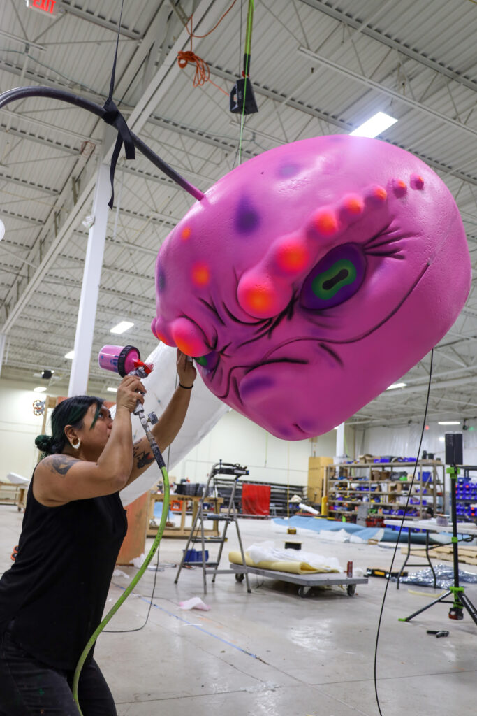 A person using a paint spray gun as they work on a large sculpture depicting a large pink-colored head of a creature.