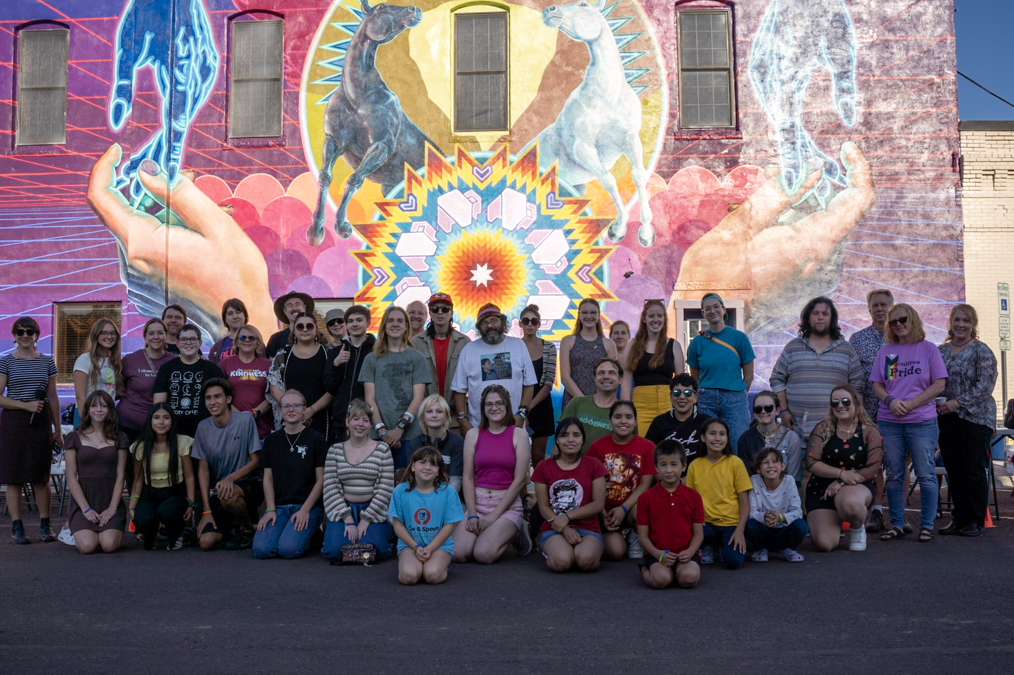 A group of people of varying ages sitting and standing in front of a brightly colored mural on a large outdoor wall.