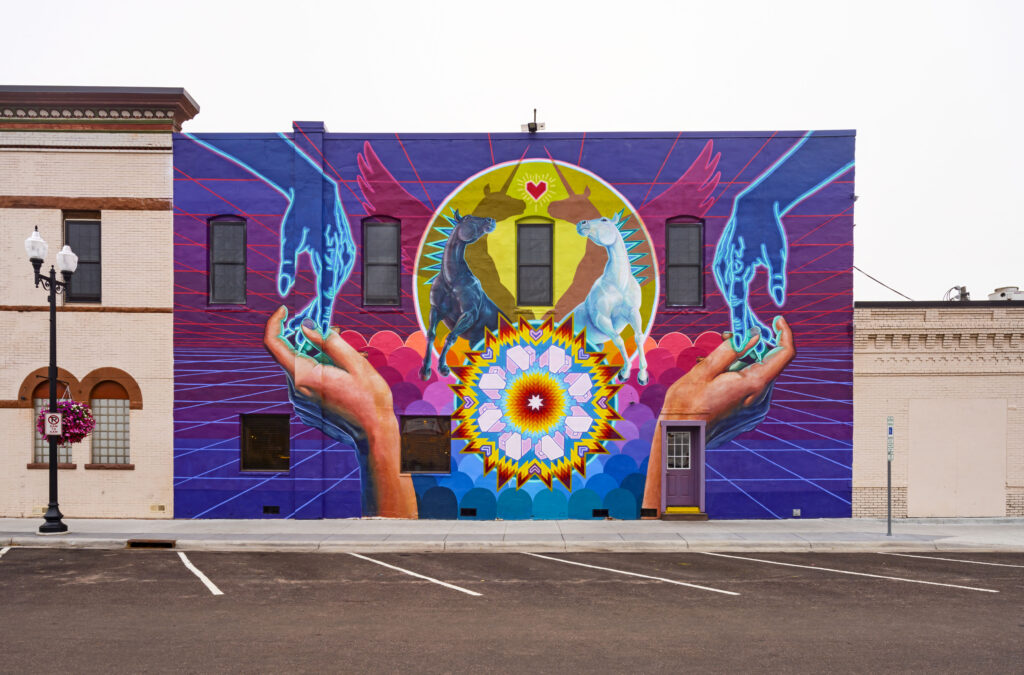 A brightly colored mural on a full facade of a two-story building. The mural depicts two pairs of hands, horses and colorful patterns.