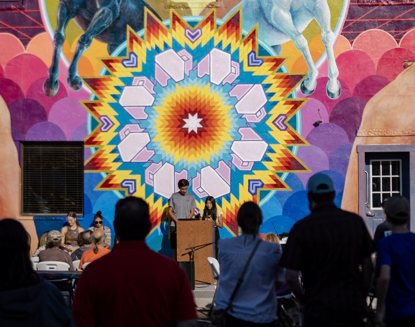 Two young people standing at a podium in front of a brightly colored mural on a large outdoor wall. There are other people sitting and standing around them.