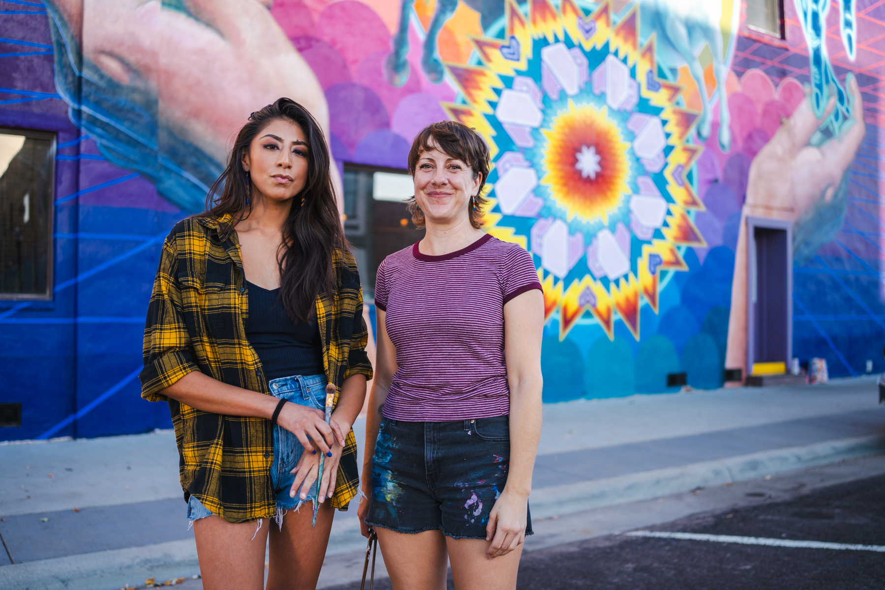 Two people standing together in front of a bright colored mural.