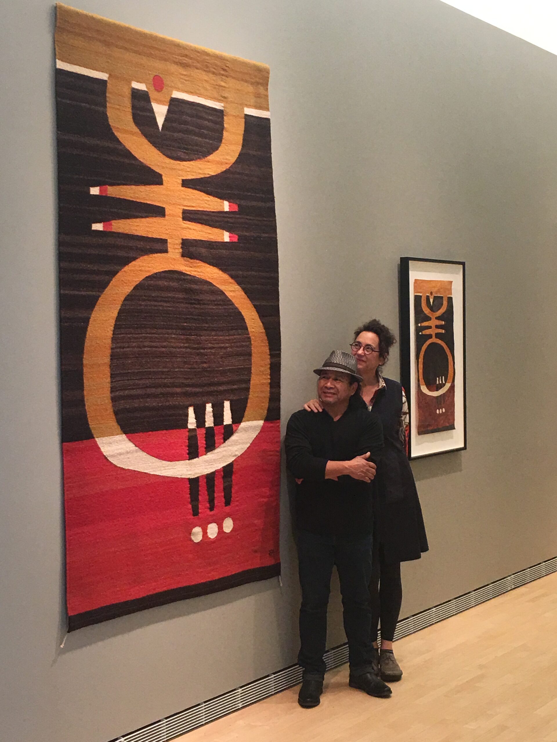 Two people stand by a hung woven textile work that looks similar to a work on paper mounted behind them on a wall.