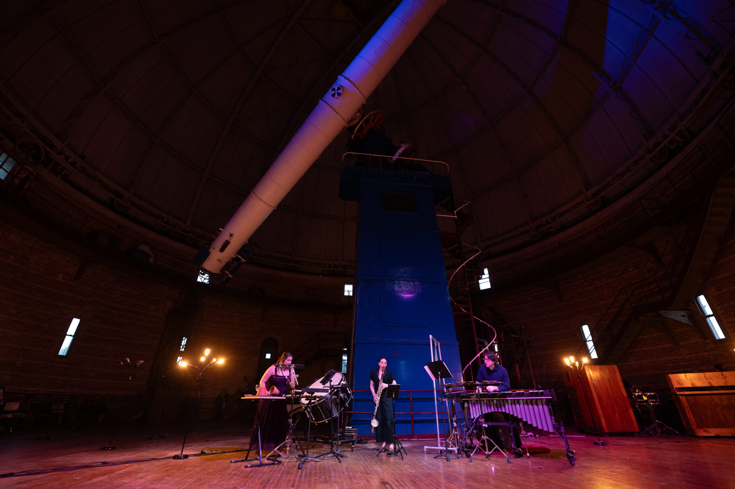 Three musicians with various instruments perform under a large refracting telescope. The observatory room is dimly lit.