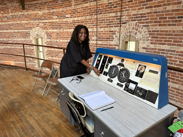 A person of dark skin tone with their dark hair in locs stands by a mechanical table inside an observatory.