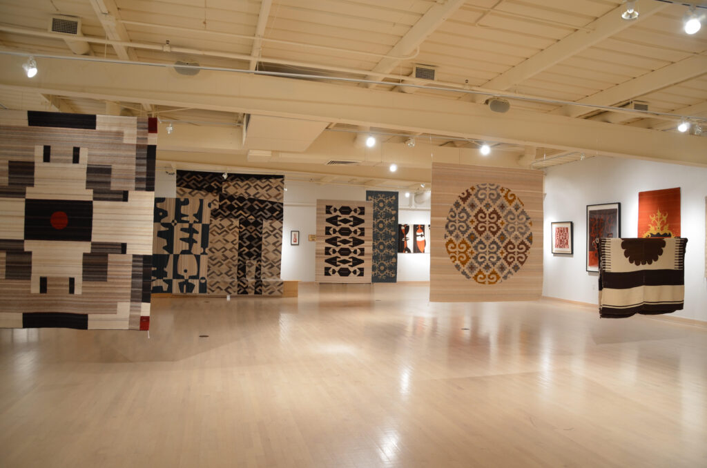 A large open room with woven textiles hanging from the ceiling and other works mounted on the walls.
