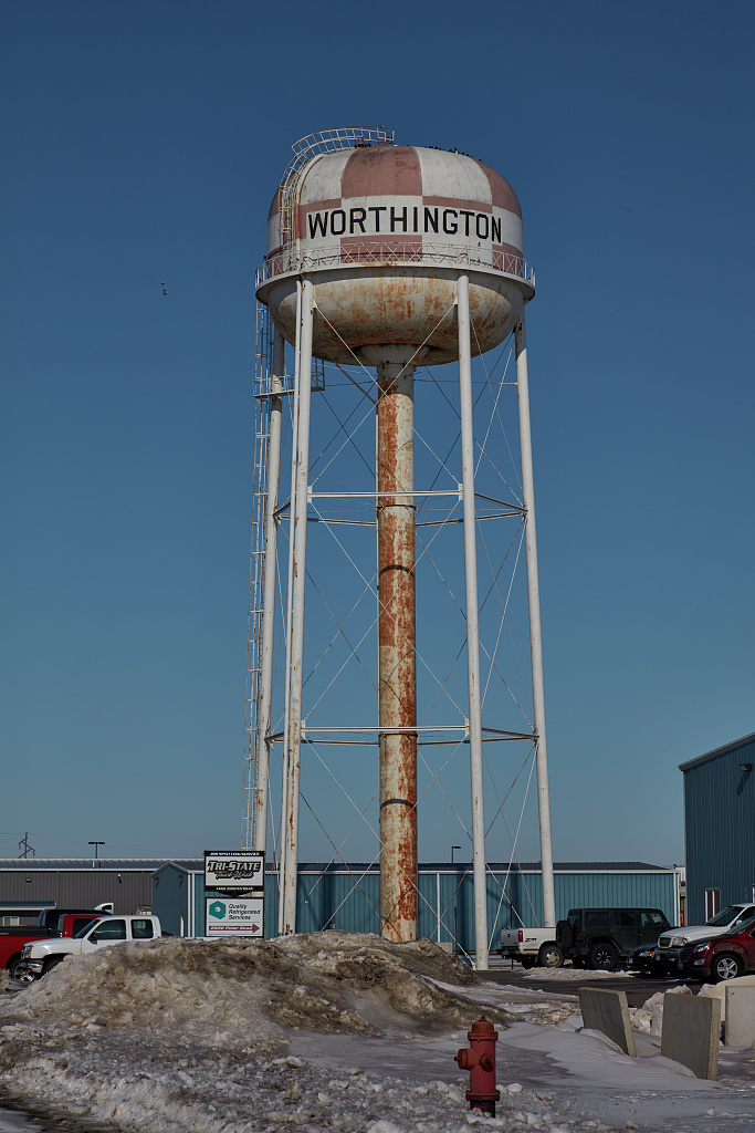 A water tower with some rusting and a faded red and white checkered pattern on the tank. In clean, black all-cap lettering, there's a painted sign on the tank that reads "Worthington'