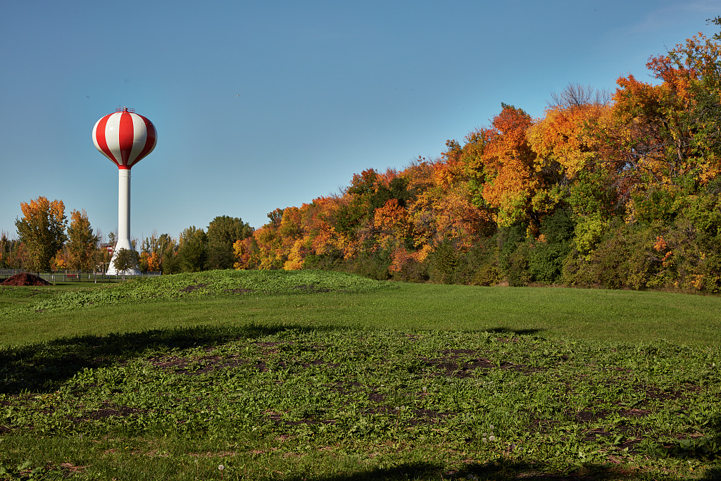 A landscape view of a field with a line of trees in fall colors to one side and a red and white striped hot air balloon-inspired water tower at the other end.