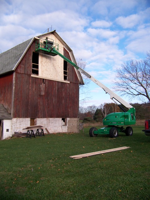 A construction lift parked by a tall wooden barn as a person works on the barn siding.