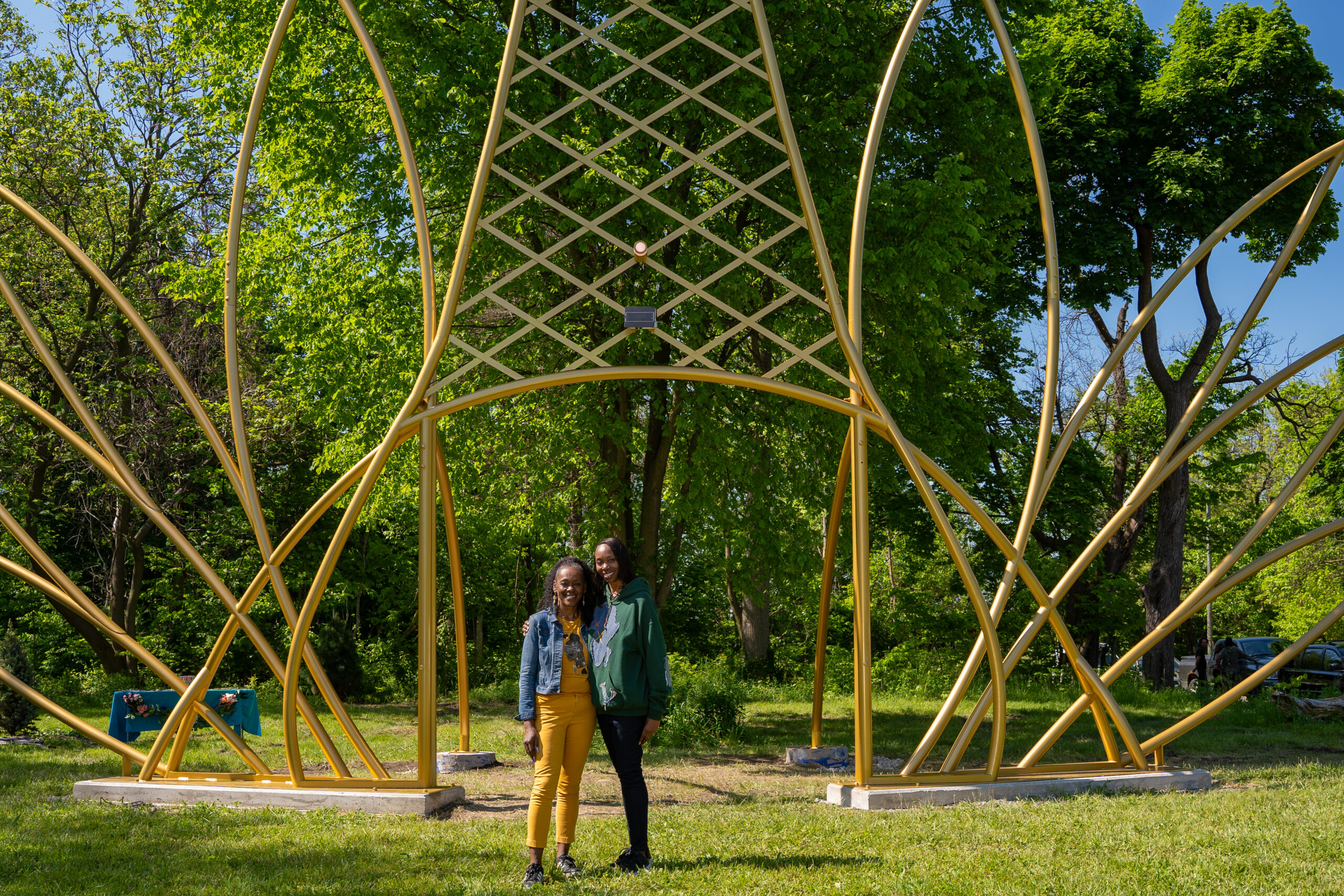 Two people at a park stand in front of a large sculptural gold-colored artwork that looks like a enlarged crown.