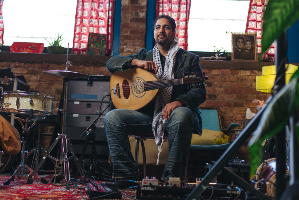 A man holds an oud on his lap in a funky looking practice space