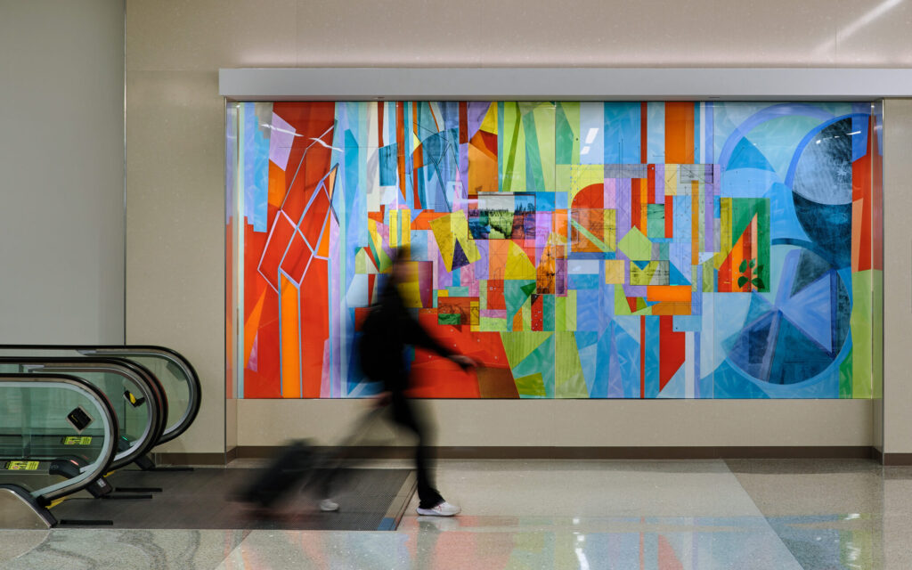 A brightly colored art installation with geometric patterns and lines. The artwork is mounted on a large wall by a set of escalators.