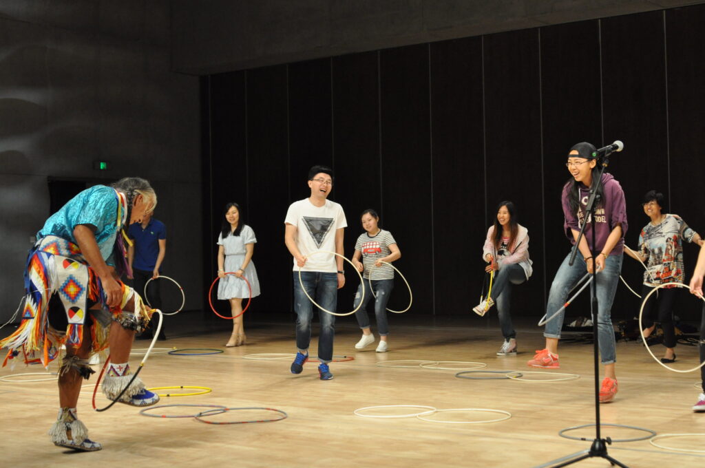 A native dancer in regalia jumps through a hoop with a group of Chinese young people