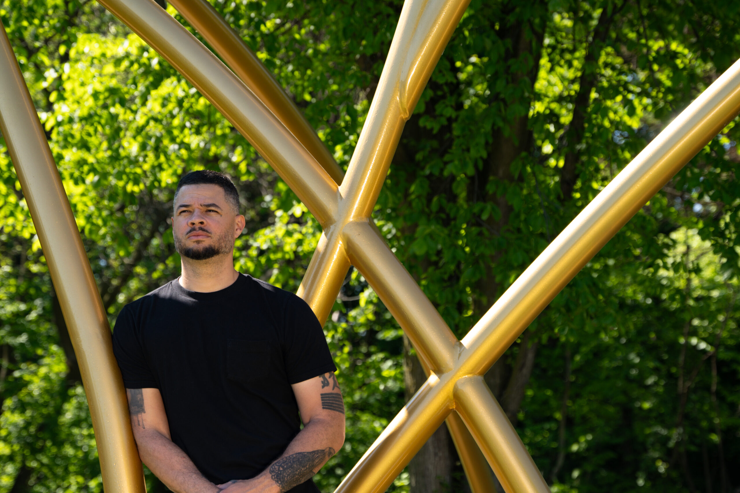 A person of medium light skin tone with short dark hair wearing a black tshirt leans on a gold-colored sculpture.