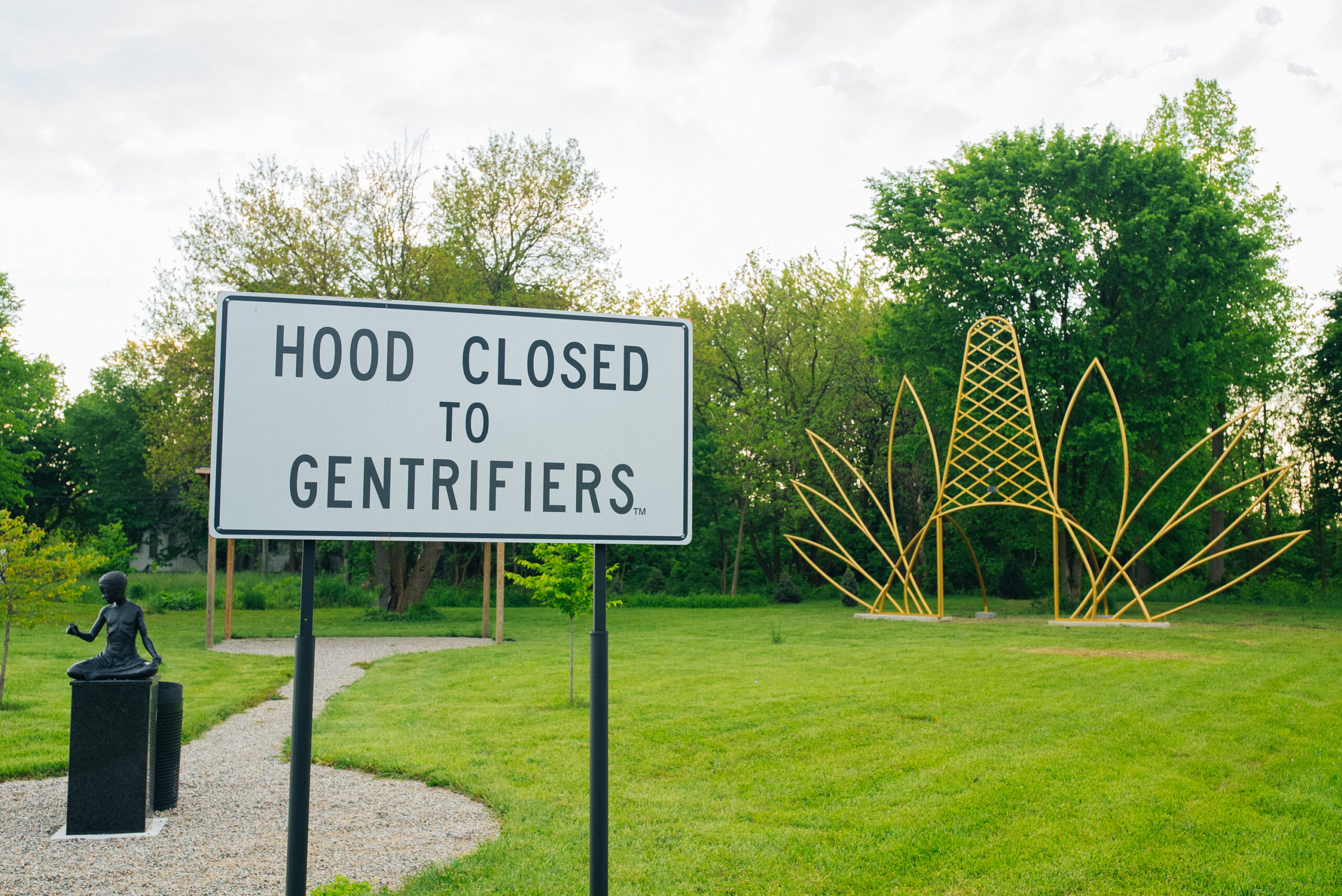 A park with different sculptural artworks including a large text-based sign that reads "Hood closed to gentrifiers"