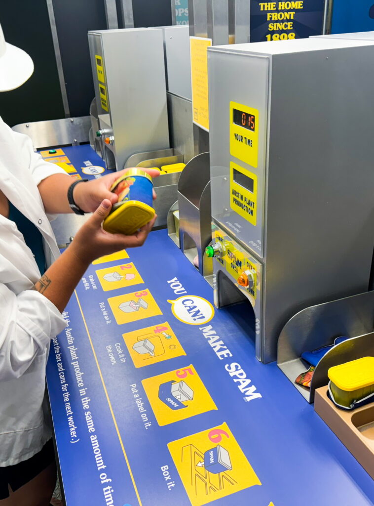 A person sliding a label onto a toy SPAM can, at a game with signage reading, "You CAN make SPAM!"