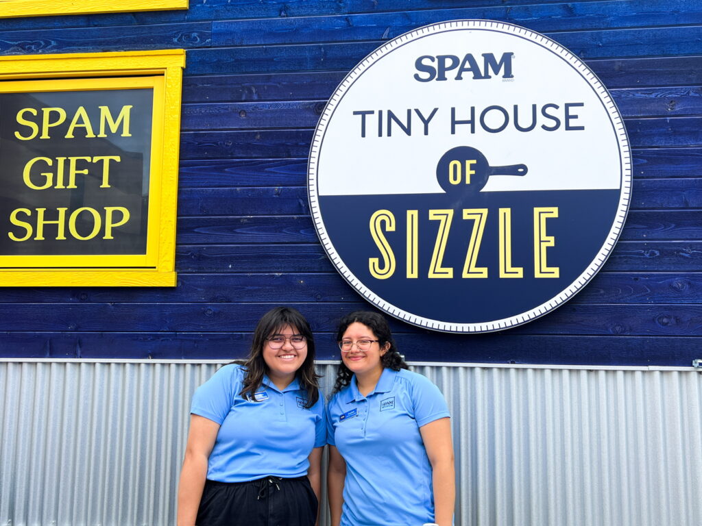 Two people in blue shirts stand in front of a tiny house with signs reading "SPAM Gift Shop" and "SPAM Tiny House of Sizzle"