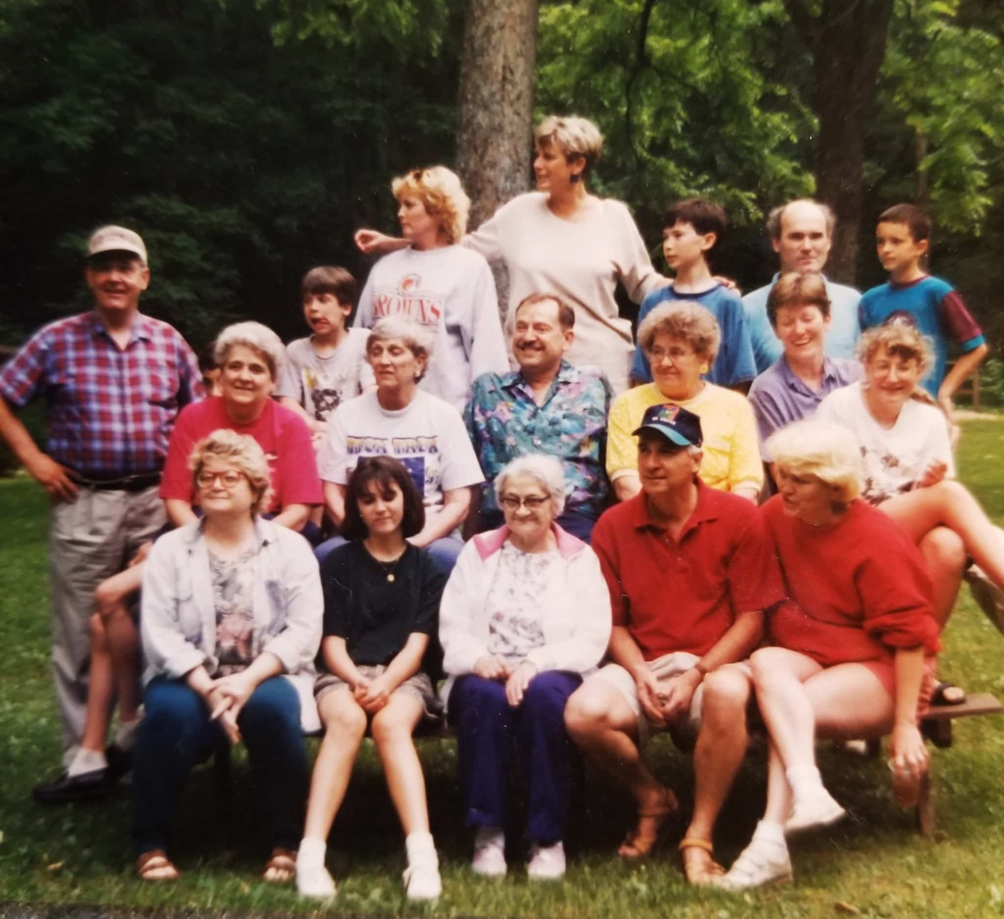 A group of about 20 people of varying ages are gathered together in rows. Some look at the camera, other beckon for those out of frame. They are seated on a bench and standing behind the bench in front of a dark forest.