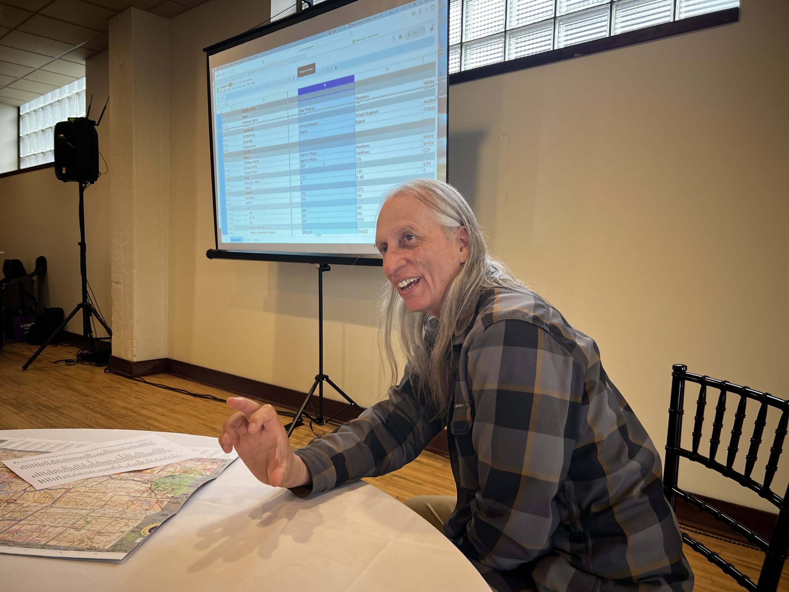 A person with long grey hair leans over a table smiling.