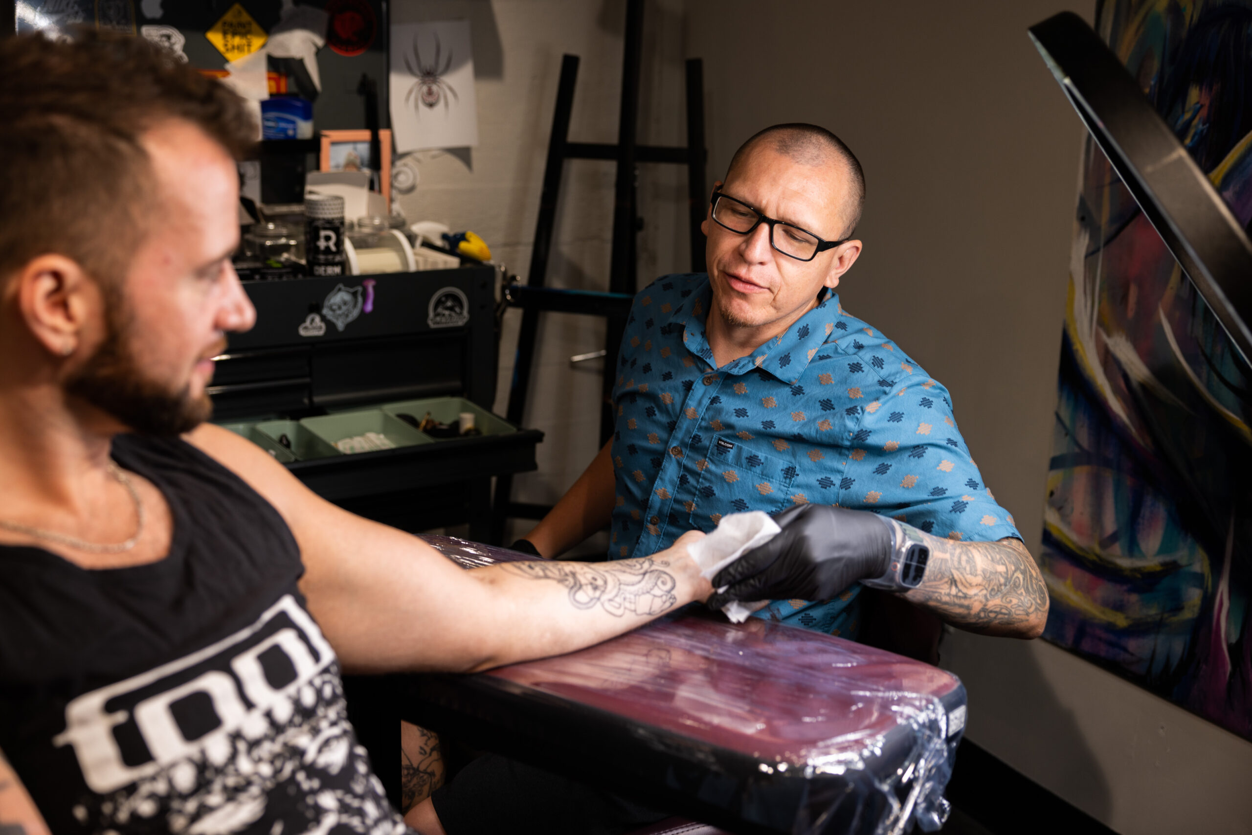 A person of medium light skin tone with a buzzed head is wiping another person's arm as they tattoo on them.