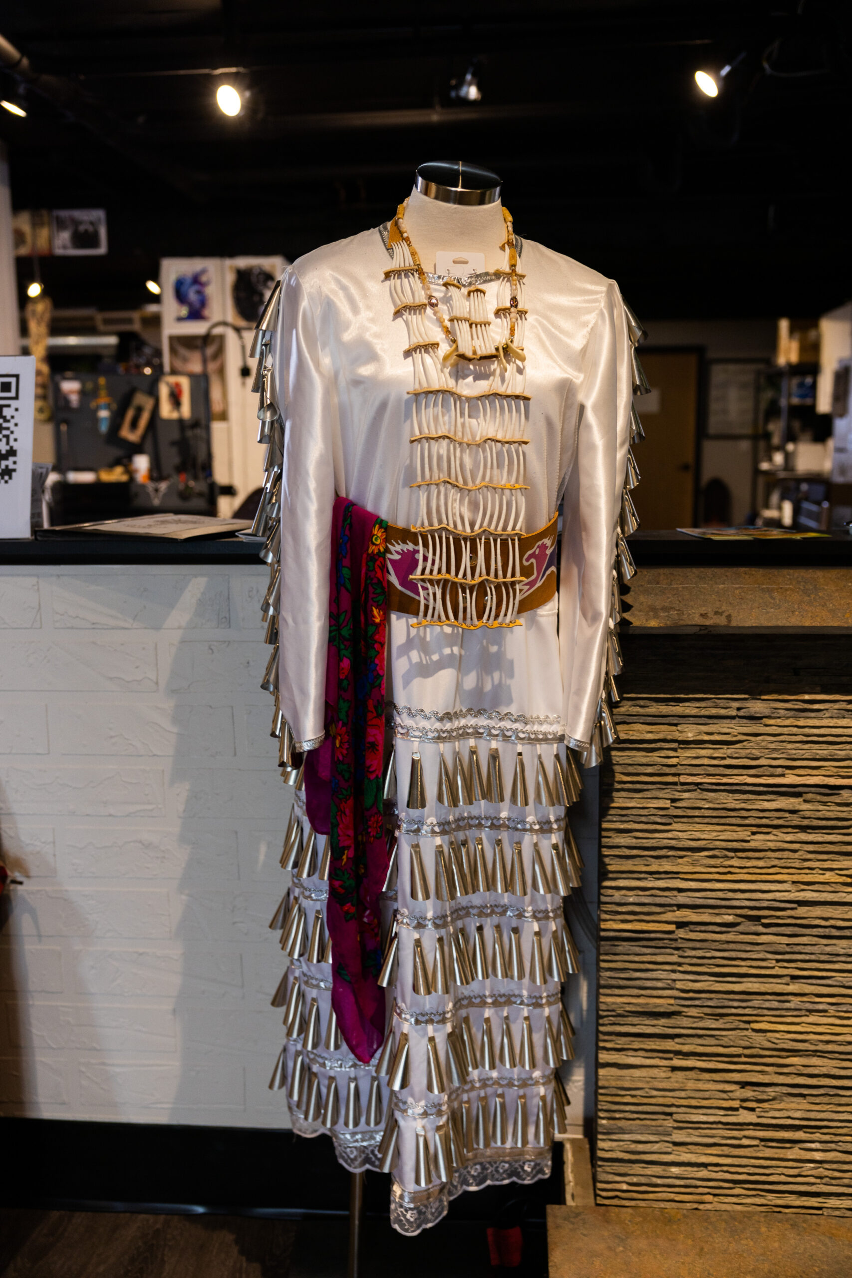 A white dress on display with the skirt and sleeves embellished with small silver jingle bells. There are other jewelry and accessories on display as well.